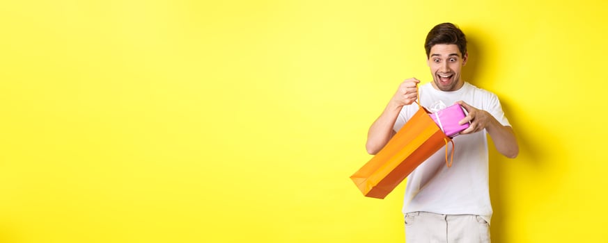 Concept of holidays and celebration. Young man looking surprised as take out gift from shopping bag, standing over yellow background photo