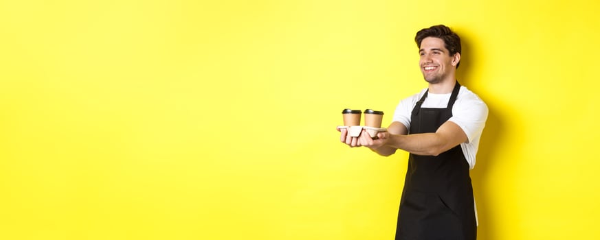 Friendly barista in black apron giving takeaway order, holding two cups of coffee and smiling, standing over yellow background photo