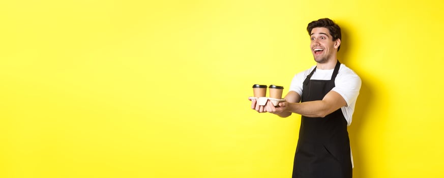 Friendly barista in black apron giving takeaway order, holding two cups of coffee and smiling, standing over yellow background photo