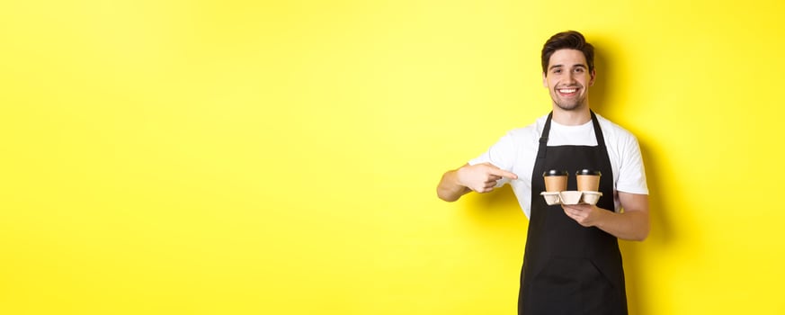 Handsome barista holding two cups of takeaway coffee, pointing finger at drinks and smiling, standing in black apron against yellow background photo