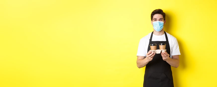 Concept of covid-19, cafe and social distancing. Barista in medical mask serving coffee in takeaway cups, standing in black apron against yellow background photo