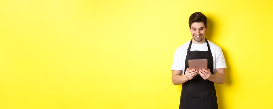 Seller in black apron looking at digital tablet screen, smiling pleased, standing over yellow background photo