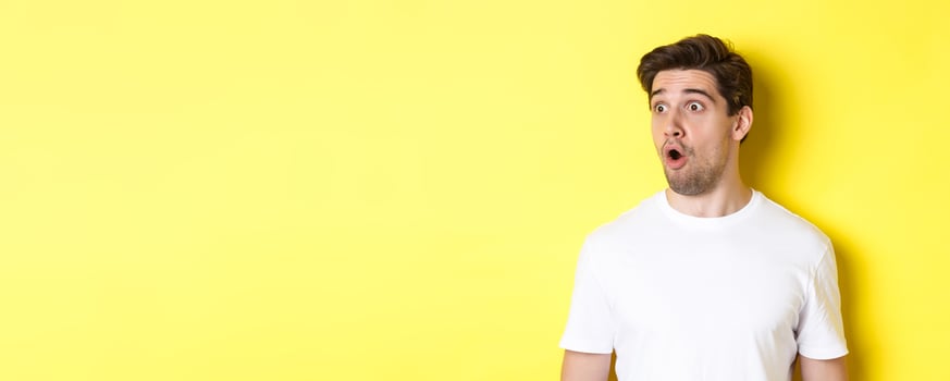 Close-up of impressed man looking left, gasping amazed, standing in white t-shirt against yellow background photo