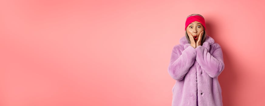 Fashion concept. Trendy old asian woman looking surprised at camera. Mature female in purple coat and headband looking amazed at camera, checking out promotion on pink background photo