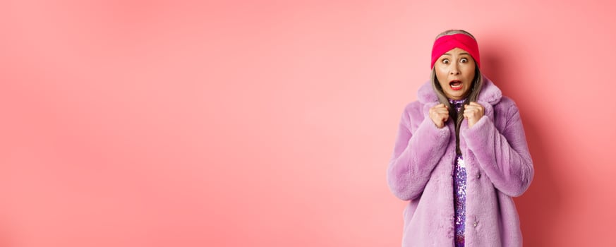 Fashion and shopping concept. Scared elderly woman scream and staring startled at camera, jumping from fear, wearing purple winter coat and headband, pink background photo
