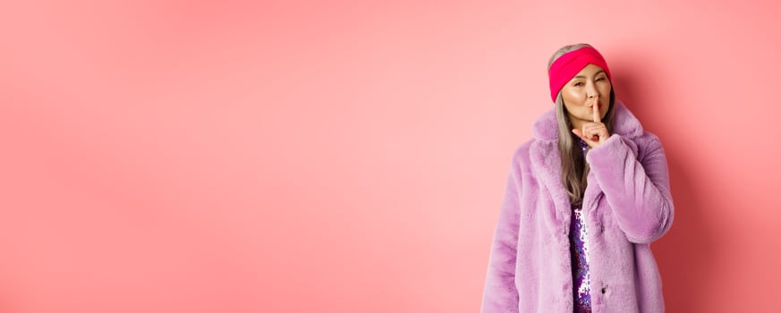 Fashion and shopping concept. Modern chinese senior woman shushing, telling a secret and looking cunning at camera, standing in trendy fake fur coat and headband on pink background photo