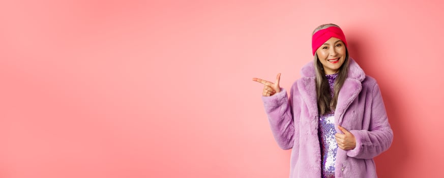Fashion and shopping concept. Stylish senior asian woman pointing finger left at promotion deal, smiling at camera, suggesting special offer, standing against pink background photo