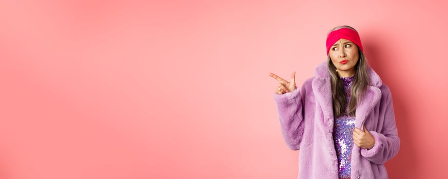 Fashion and shopping concept. Skeptical middle-aged asian woman in purple fur coat looking and pointing finger left with displeased, sad expression, standing over pink background photo