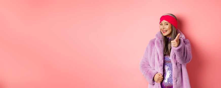 Fashion and shopping concept. Happy asian mature woman in stylish fake-fur coat pointing finger at camera, we need you gesture, standing over pin background photo