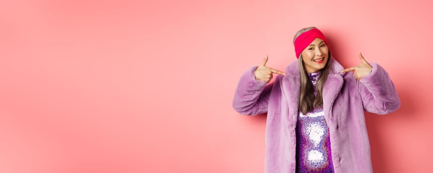 Fashion and shopping concept. Cool and trendy asian senior woman pointing fingers at herself, dressed in stylish purple fake-fur coat and dress, smiling happy, pink background photo