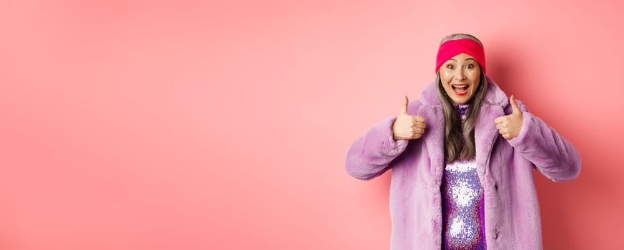Fashion and shopping concept. Cheerful asian old woman showing thumbs-up in approval, like and recommend promotion, standing in stylish purple fur coat, pink background photo