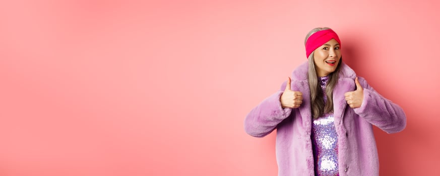 Fashion and shopping concept. Stylish asian granny saying yes. Trendy senior korean woman in winter fake-fur coat show thumbs-up in approval, praising good product, pink background photo