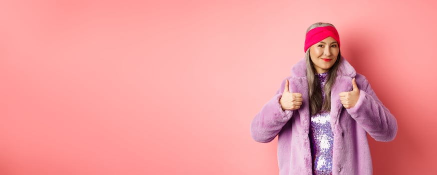 Fashion and shopping concept. Asian fashionable lady in winter coat showing thumbs-up, smiling and looking satisfied, recommending company promo, pink background photo