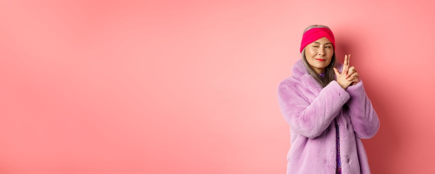 Fashion and shopping concept. Cool and stylish asian senior lady in purple fake fur coat, making finger gun gesture and looking left with sassy smile, acting like secret agent, pink background photo