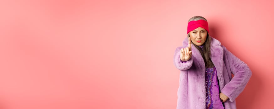 Fashion and shopping concept. Serious asian senior woman showing not so fast gesture, shaking extended finger to stop or warn you, looking determined at camera, wearing stylish purple clothes photo
