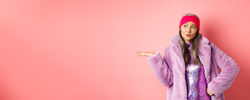 Fashion and shopping concept. Stylish asian senior woman in purple winter outfit demonstrating product on hand. Cool old lady in trendy clothes holding something on palm, pink background photo