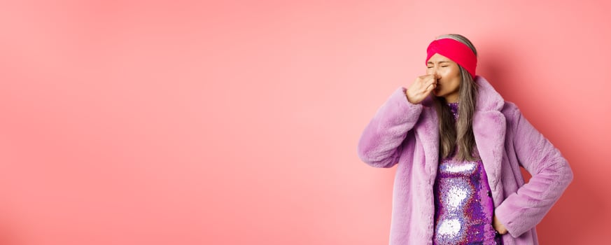 Portrait of stylish old asian woman in fashionable purple coat and dress, shut nose from bad smell, express disgust and aversion as something sink awfully, standing over pink background photo