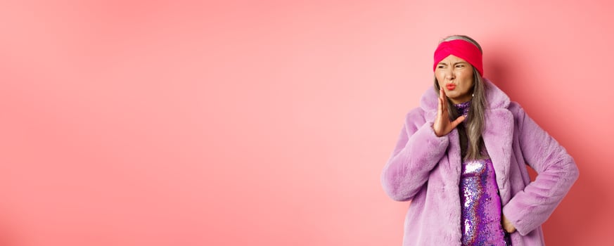 Fashion and shopping concept. Stylish asian middle-aged woman in fashionable winter clothes smell something bad, waving hand near nose from disgusting stink, pink background photo