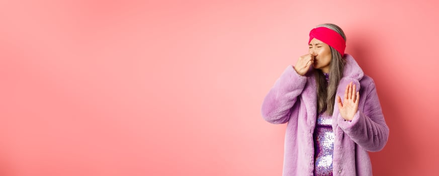 Disgusted asian mature woman in trendy purple winter coat and dress, shut nose and showing stop, refusal gesture, smell something disgusting, standing over pink background photo