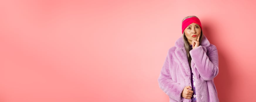 Funky old asian woman looking thoughtful and perplexed, standing in fake fur coat and thinking, making decision, standing on pink background photo