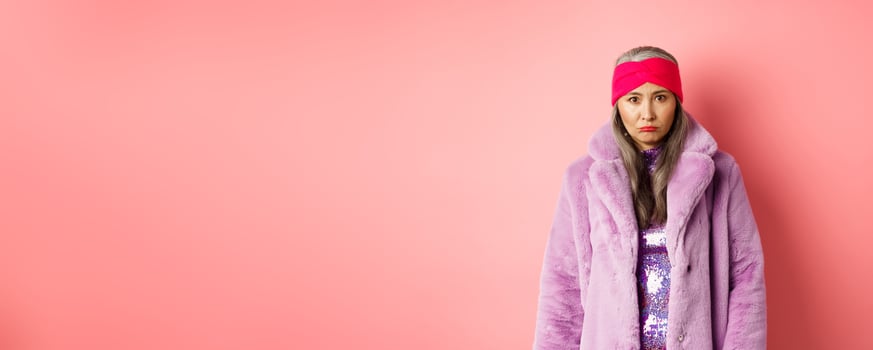 Sad and gloomy stylish asian senior woman looking upset at camera, sulking and feeling distressed, standing in purple faux fur coat against pink background photo