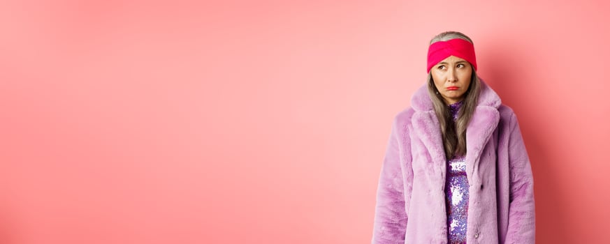 Sad and indecisive asian senior woman in fashionable faux fur coat looking left and sulking upset, standing against pink background photo