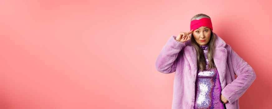 Stylish asian mature woman scolding you, staring at camera annoyed and confused, pointing finger at head, are you stupid gesture, standing over pink background photo