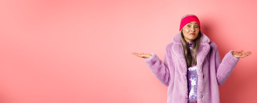 Fashionable and funky asian old woman shrugging shoulders, looking confused and clueless, standing in stylish purple coat and headband, pink background photo
