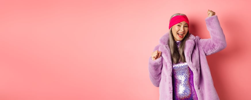 Shopping concept. Cheerful senior asian woman dancing and having fun, wearing stylish purple faux fur coat and dress, winning prize and smiling, standing over pink background photo
