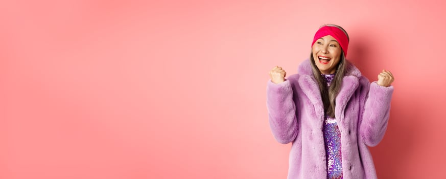 Asian senior woman celebrating, cheering and winning, making fist pump signs in triumph, achieve goal and smiling satisfied, standing in stylish faux fur coat, pink background photo