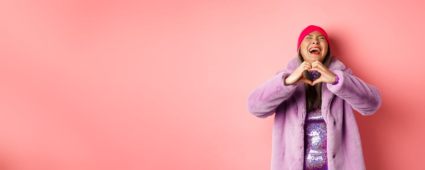 Valentines day and shopping concept. Happy asian woman in stylish outfit showing heart sign and laughing carefree, standing in faux fur against pink background photo