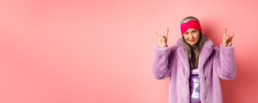 Funny and cool asian senior woman showing rock n roll gesture, looking sassy, standing over pink background photo