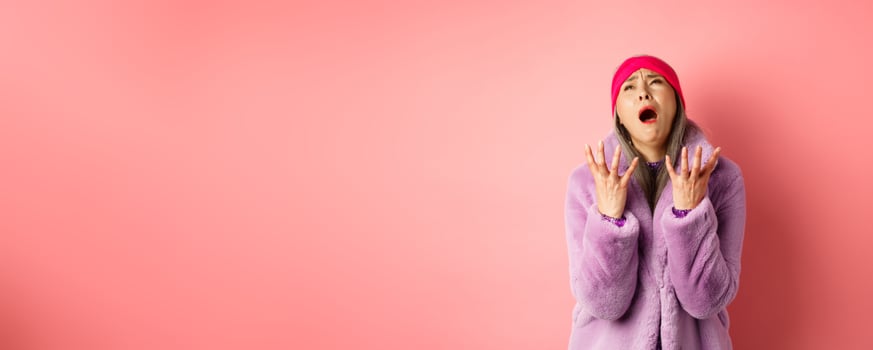 Miserable asian senior woman in stylish purple faux fur coat, pleading god and asking why, shaking hands and looking up desperate, standing over pink background photo