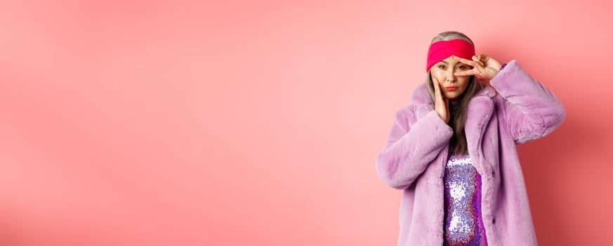 Fashion and shopping. Beautiful asian senior woman in stylish faux fur coat and headband, making peace sign on face and looking sassy and confident at camera, pink background photo