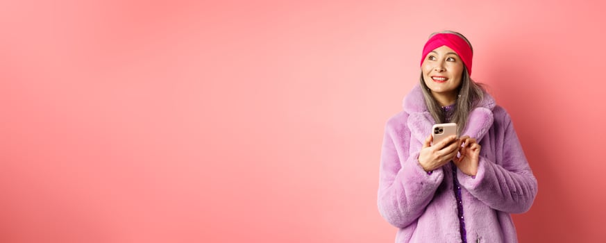 Online shopping and fashion concept. Stylish asian senior woman using smartphone, looking aside with thoughtful face, standing against pink background photo