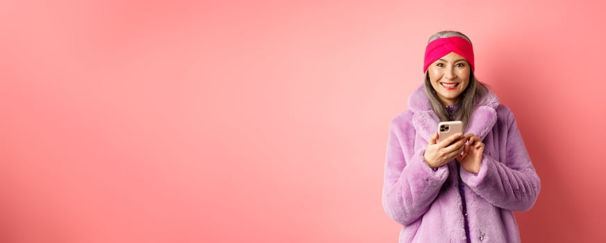 Online shopping and fashion concept. Smiling asian senior lady in winter coat and headband order in internet, holding mobile phone and looking happy, pink background photo