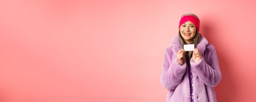 Shopping and fashion concept. Beautiful asian middle-aged woman showing plastic credit card and smiling happy in stylish clothes, pink background photo