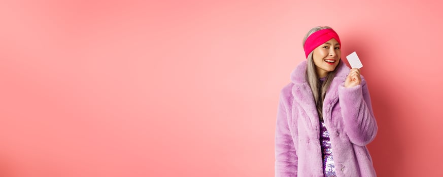 Shopping and fashion concept. Stylish asian senior woman in trendy clothes showing plastic credit card, smiling sassy, standing over pink background photo