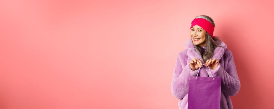 Shopping and fashion concept. Fashionable gradmother bought present and smiling, holding purple bag in hands, standing over pink background photo
