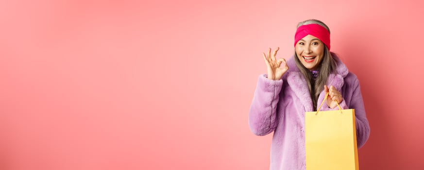 Shopping and fashion. Happy asian senior woman in stylish clothes holding paper bag from store, showing OK sign, recommending shop, pink background photo
