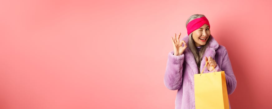 Stylish senior asian woman recommending store, holding shopping bag and winking with OK sign, smiling satisfied, standing over pink background photo