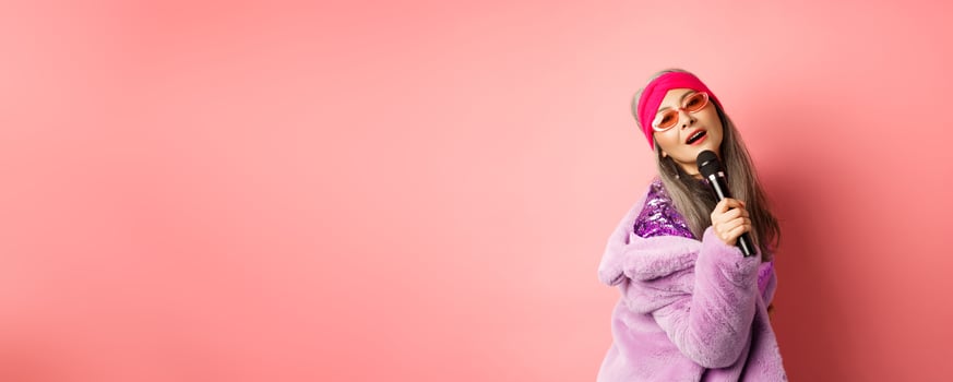 Stylish and sassy asian mature woman perform song on stage, holding mic and singing karaoke, wearing trendy heart-shaped sunglasses and faux fur coat, pink background photo