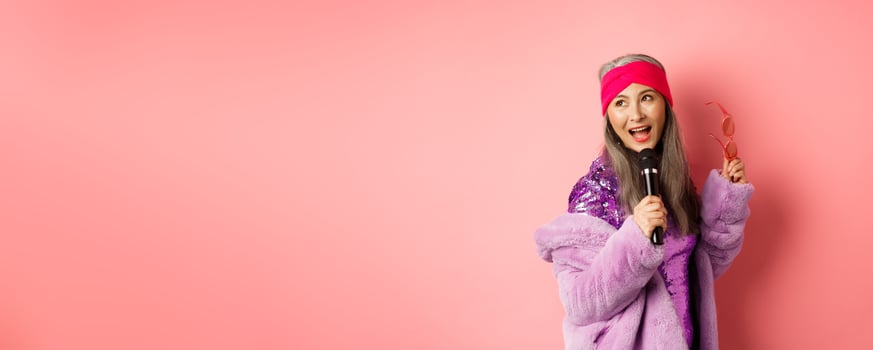 Gorgeous asian senior woman singing karaoke in mic, perform song and looking happy, standing over pink background photo
