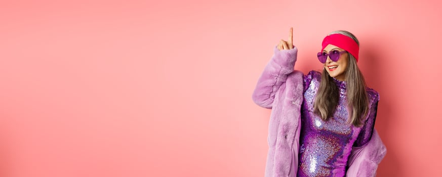 Asian old woman in fashionable glittering dress and purple faux fur coat dancing disco, pointing finger up and having fun, pink background photo