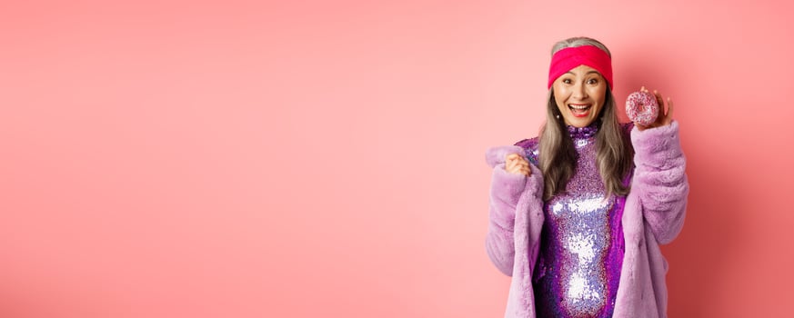 Cheerful asian senior woman with grey hair, wearing party glitter dress, showing delicious donut and smiling happy, eating sweet food, standing over pink background photo
