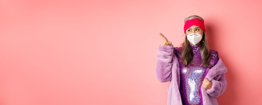Covid and fashion concept. Stylish asian senior woman checking out special deal, wearing disco dress and face mask, pointing finger left at copy space, pink background photo