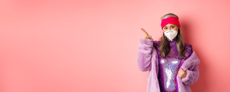 Covid and fashion concept. Fashionable asian mature woman in face mask and disco dress pointing finger upper left corner. Old lady in glittering dress showing advertisement photo