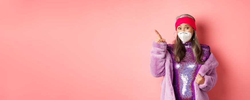 Covid and fashion concept. Amazed asian senior woman in glittering dress and face mask, pointing finger upper left corner, showing promo offer, pink background photo