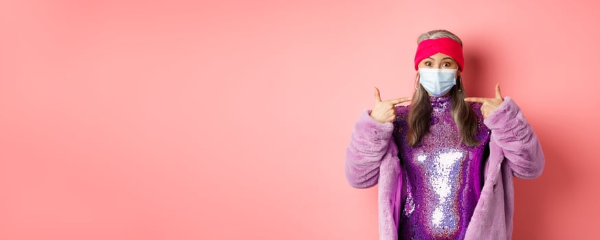 Covid-19, social distancing and fashion concept. Asian edlerly woman in stylish party outfit and respirator, pointing fingers at face mask, asking to follow coronavirus quarantine rules photo
