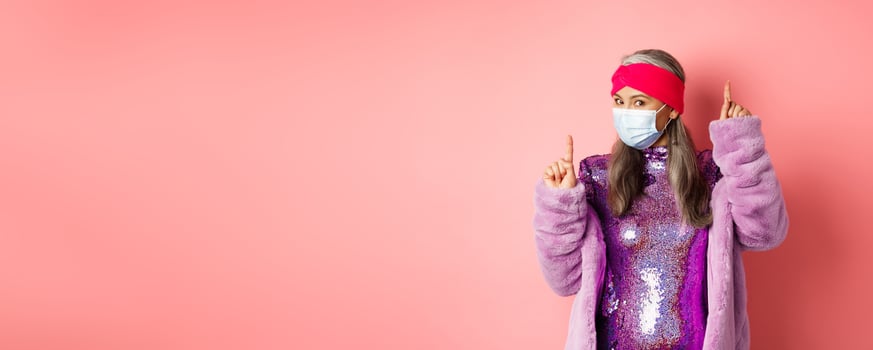 Covid-19, social distancing and fashion concept. Cheerful asian senior woman likes to party, dancing in respirator and shiny purple dress, standing over pink background photo